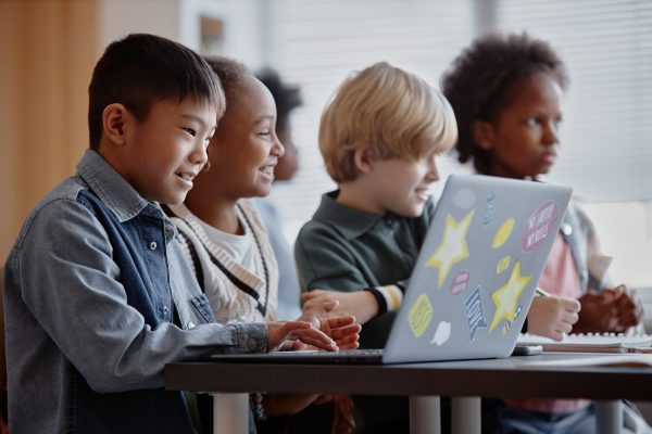 Side view of diverse group of happy primary students sitting in row using laptop during lesson